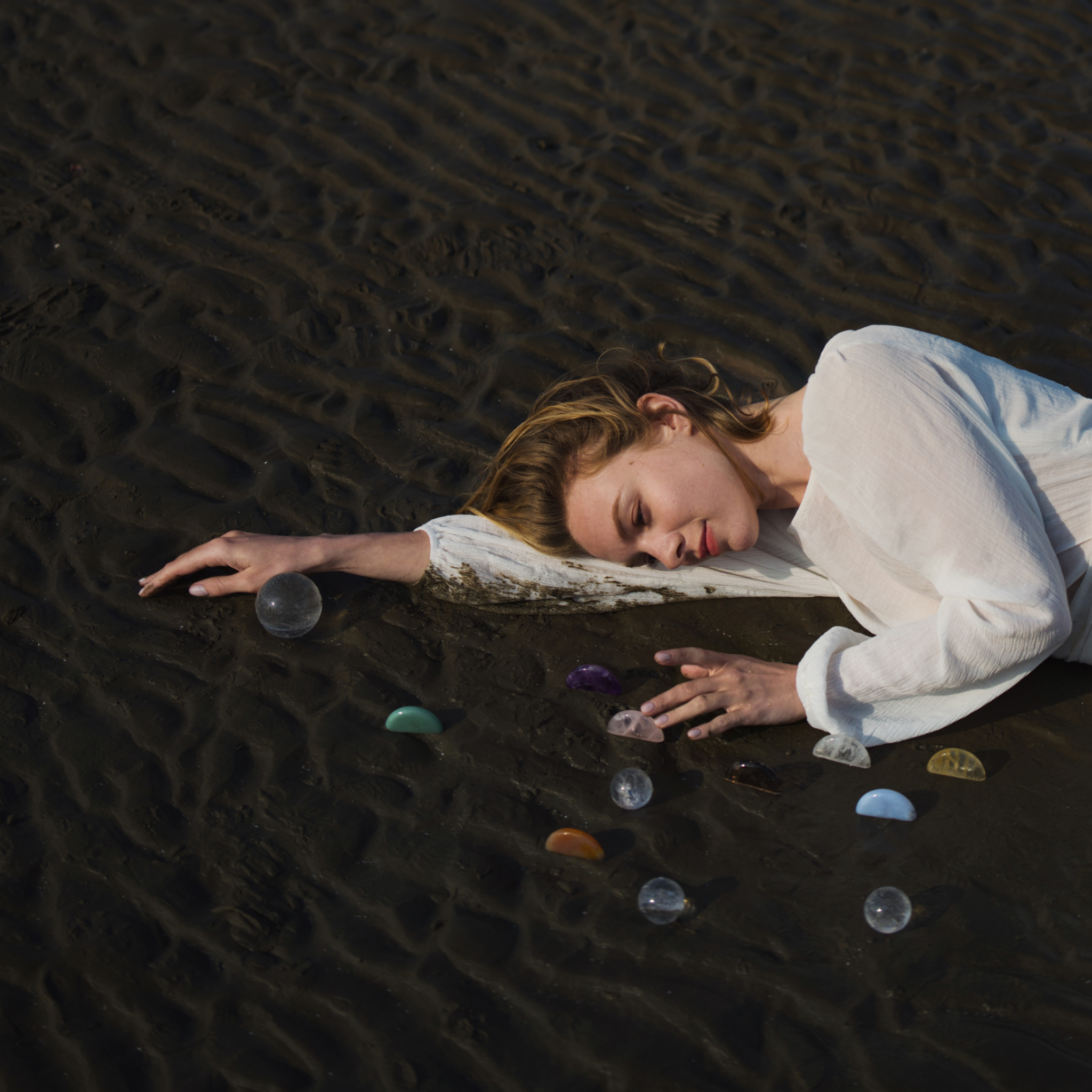 Woman in White Dress Lying on Beach Surrounded by Eight Natural Healing Crystals for Energy and Chakra Balancing