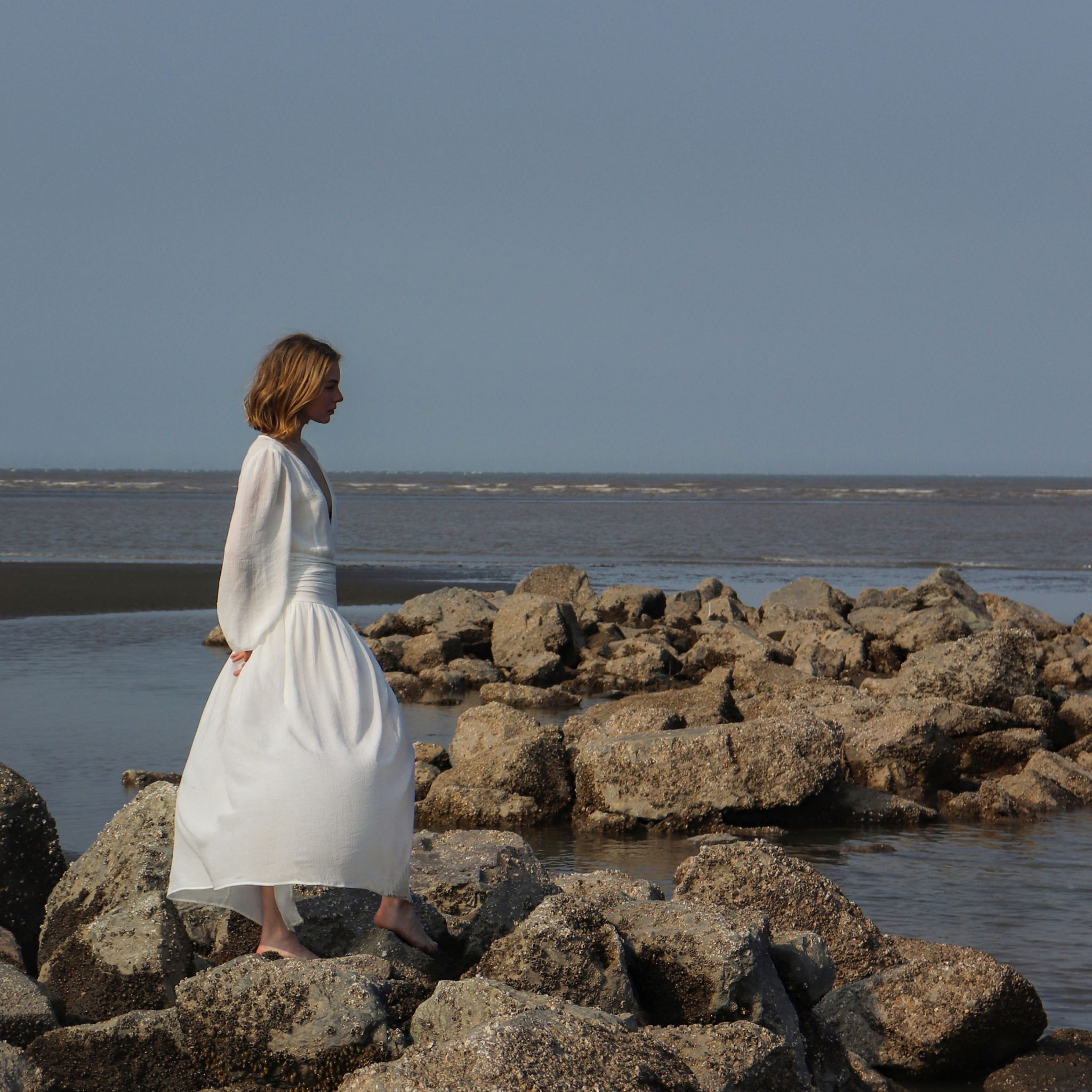 Woman in a white dress standing on rocky beach with ocean in the background