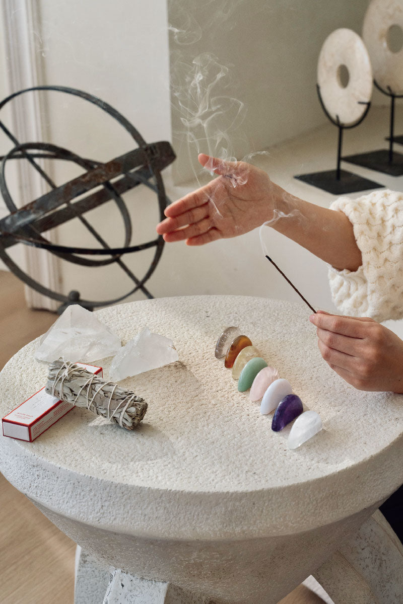 Hand waving sacred White Sage smoke over chakra crystals for botanical purification, a ritual to clear stagnant energy