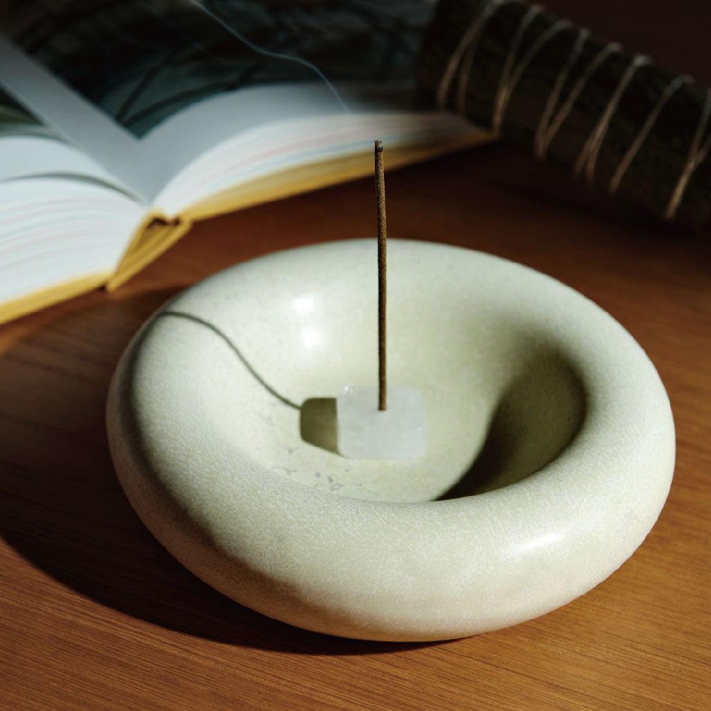 Burning incense stick standing in a clear crystal holder placed inside a ceramic ritual bowl. Warm wooden setting with an open book and cedar bundle in the background for meditation