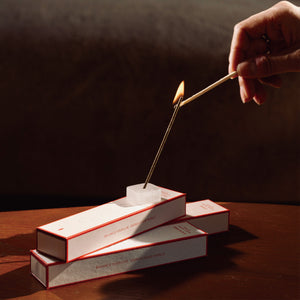 Hand lighting an incense stick placed in a real crystal holder atop stacked Orblink incense boxes. Warm atmospheric lighting for a purification ritual.